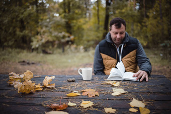 年轻的男人。坐在秋天公园木表格饮料茶读取书温暖的黑暗秋天大气软焦点模糊复制空间