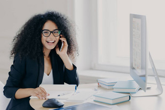 乐观女非洲式发型美国企业家卷曲的发型持有移动电话享受电话谈话的同事作品电脑提出了工作场所记事本书