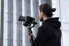 年轻的专业电视录像制作人持有专业相机轴健身球稳定剂为设备帮助使高质量视频摇晃摄影师穿白色连帽衫使视频