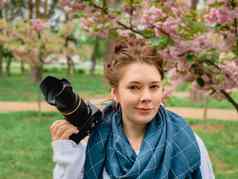 年轻的快乐微笑红发女人白色毛衣蓝色的围巾持有手相机开花粉红色的樱花树公园城市基辅乌克兰