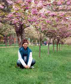 年轻的快乐微笑女人牛仔裤裤子白色毛衣蓝色的围巾坐在草开花粉红色的樱花树公园城市基辅乌克兰