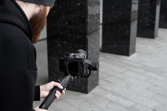 有胡子的专业电视录像制作人黑色的连帽衫持有专业相机轴健身球稳定剂导演使伟大的视频专业电影相机摄影师