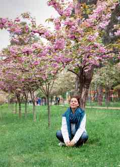 年轻的快乐微笑女人牛仔裤裤子白色毛衣蓝色的围巾坐在草开花粉红色的樱花树公园城市基辅乌克兰
