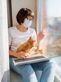 女人医疗面具远程作品首页坐在窗口窗台上移动PC可爱的姜猫膝盖封锁检疫冠状病毒科维德隔离首页