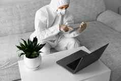 男人。保护白色西装医疗面具工作首页沙发移动PC平板电脑冠状病毒疫情远程工作流感大流行保持首页科维德检疫概念