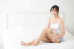 美丽的年轻的亚洲女人坐着床上抚摸腿软光滑的皮肤卧室女孩应用触摸身体奶油乳液治疗护理健康的健康概念