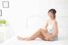 美丽的年轻的亚洲女人坐着床上抚摸腿软光滑的皮肤卧室女孩应用触摸身体奶油乳液治疗护理健康的健康概念