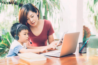 年轻的亚洲妈妈。儿子移动PC电脑平板电脑研究学习首页男孩穿耳机电子学习距离老师妈妈支持孩子教育概念