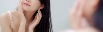 美丽的年轻的亚洲女人镜子检查脸皮肤护理化妆品卫生美女孩快乐清洁面部奶油乳液健康横幅网站