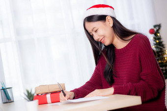 年轻的亚洲女人写作明信片圣诞节一天首页夏娃庆祝女写作消息问候卡给礼物盒子假期季节祝贺庆祝活动