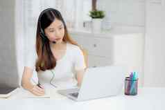 年轻的亚洲女人穿耳机研究在线电子学习桌子上女孩穿耳机学习互联网移动PC电脑首页正常的距离教育培训