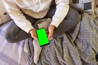 前视图年轻的女人坐着沙发上穿舒适的衣服智能手机手女人坐着交叉腿持有手机空白绿色屏幕复制空间
