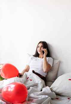 年轻的浅黑肤色的女人女人坐着醒着的床上红色的心形状的气球装饰发短信