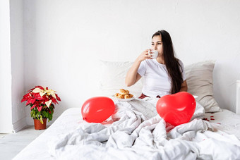 年轻的浅黑肤色的女人女人<strong>坐</strong>着醒着的床上红色的心形状的<strong>气球</strong>装饰喝咖啡吃羊角面包