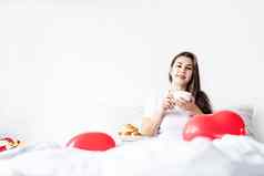 年轻的浅黑肤色的女人女人坐着醒着的床上红色的心形状的气球装饰喝咖啡吃羊角面包