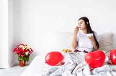 年轻的浅黑肤色的女人女人坐着醒着的床上红色的心形状的气球装饰喝咖啡吃羊角面包