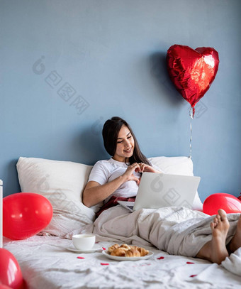 年轻的快乐浅黑肤色的女人女人<strong>坐</strong>着床上红色的心形状的<strong>气球</strong>聊天的男朋友移动PC显示心手势手