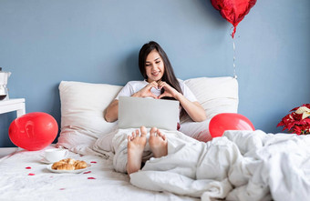 年轻的快乐浅黑肤色的女人女人坐着床上红色的心形状的气球聊天的男朋友移动PC显示心手势手