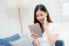 年轻的有吸引力的亚洲女人休息浏览平板电脑电脑沙发首页快乐女孩坐着沙发上放松阅读数字小工具房子沟通生活方式概念