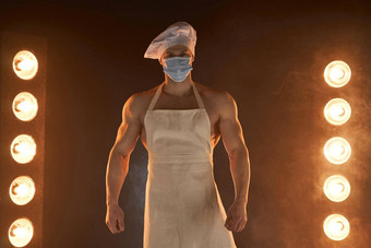 正常的概念肌肉发达的老板穿保护医疗面具围裙老板他站烟雾缭绕的背景灯照明