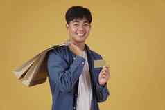 肖像快乐年轻的亚洲男人。穿着随便持有购物袋信贷卡孤立的黄色的背景