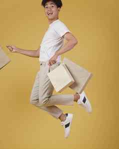 肖像快乐兴奋年轻的亚洲男人。穿着随便持有购物袋跳孤立的黄色的背景