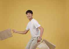 肖像快乐年轻的亚洲男人。穿着随便持有购物袋孤立的黄色的背景