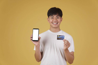 肖像快乐年轻的亚洲男人。持有智能<strong>手机</strong>信贷卡购物在线孤立的黄色的背景