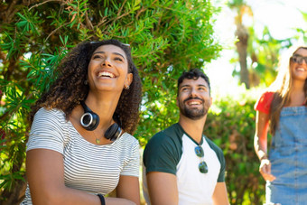 非洲式发型美国年轻的女人无线耳机坐着城市公园千禧一代朋友集团无忧无虑的赶时髦的人有趣的夏天太阳自然小镇