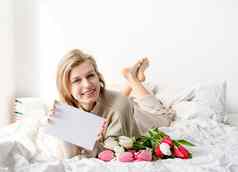 快乐女人说谎床上穿睡衣持有郁金香花花束空白卡