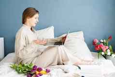 快乐女人坐着床上穿睡衣持有郁金香花花束聊天移动PC