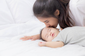 年轻的亚洲妈妈。吻脸颊婴儿女孩温柔的床上卧室妈妈爱新生儿护理妈妈。表达式孩子父女儿家庭概念