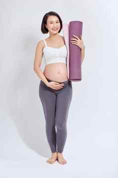 年轻的有吸引力的怀孕了女触碰肚子持有瑜伽席手准妈妈。物理练习摆姿势孤立的白色背景