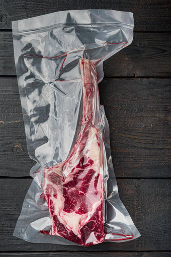新鲜的美味的生牛肉战斧牛排骨包装真空塑料包装市场黑色的木表格背景前视图平躺