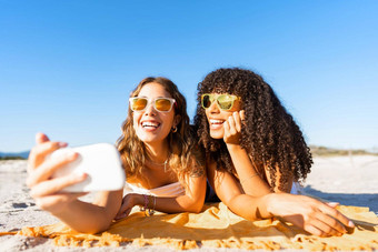 美丽的年轻的女人持有智能手机采取图片卷曲的黑色的拉美裔女朋友说谎海滩太阳镜微笑相机夏天好旅行旅游