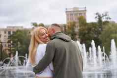 浪漫的走年轻的夫妇爱拥抱喷泉城市公园男人。窃窃私语赞美美丽的女朋友