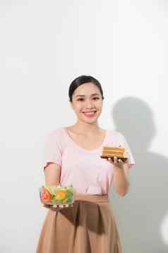 半身的肖像美丽的女人持有小蛋糕新鲜的蔬菜年轻的家庭主妇选择糖果健康的吃蛋糕沙拉孤立的白色背景