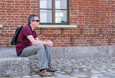 成人养老金领取者背包坐在休息背景红色的砖建筑