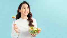肖像快乐好玩的女孩吃新鲜的沙拉碗眨眼孤立的蓝色的背景