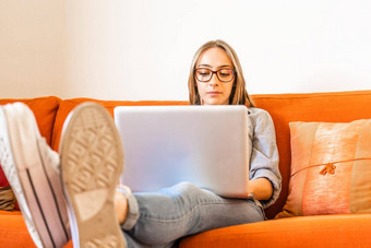 可爱的金发女郎女孩眼镜坐着首页沙发集中研究工作移动PC电脑脚休息表格年轻的企业家女人工作首页笔记本无线网络