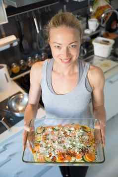 微笑年轻的健康的女人持有自豪地显示玻璃烘焙行素食者菜成分把烤箱
