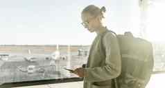 年轻的女人站机场盖茨窗口持有手机手穿旅行背包走休息室区域女旅行者搜索在线地图机场