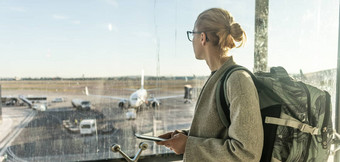 年轻的女人站机场盖茨窗口持有手机手穿旅行背包走休息室区域女旅行者搜索在线地图机场