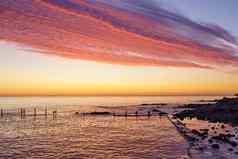 橙色红色的天空华丽的云被遗弃的海码头黎明日落水反射岩石