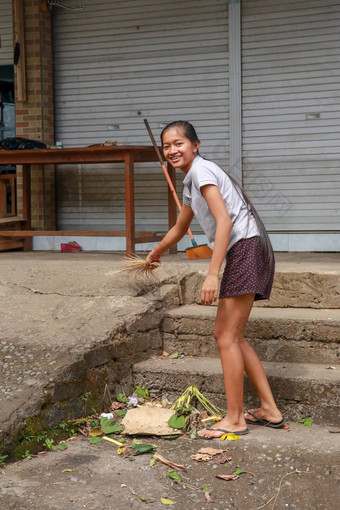 女人清洁服务<strong>扫</strong>帚坚持簸箕清洁路巴厘岛的女孩清洁工垃圾干叶子前面<strong>房子</strong>巴厘岛印尼