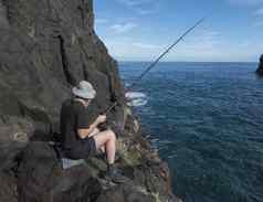回来一边年轻的男人。渔民他坐着边缘熔岩岩石悬崖钓鱼杆海岸大西洋海洋棕榈岛西班牙