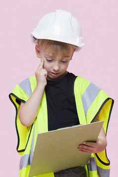 肖像深思熟虑的年轻的男孩穿着建设工人剪贴板粉红色的背景