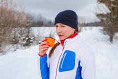 年轻的女人冬天体育运动夹克喝热茶橙色