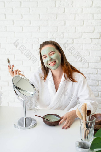 年轻的微笑高加索人女人穿浴袍appplying粘土脸面具镜子使有趣的脸