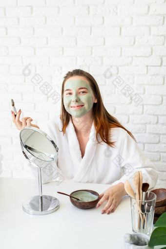 年轻的微笑高加索人女人穿浴袍appplying粘土脸面具镜子